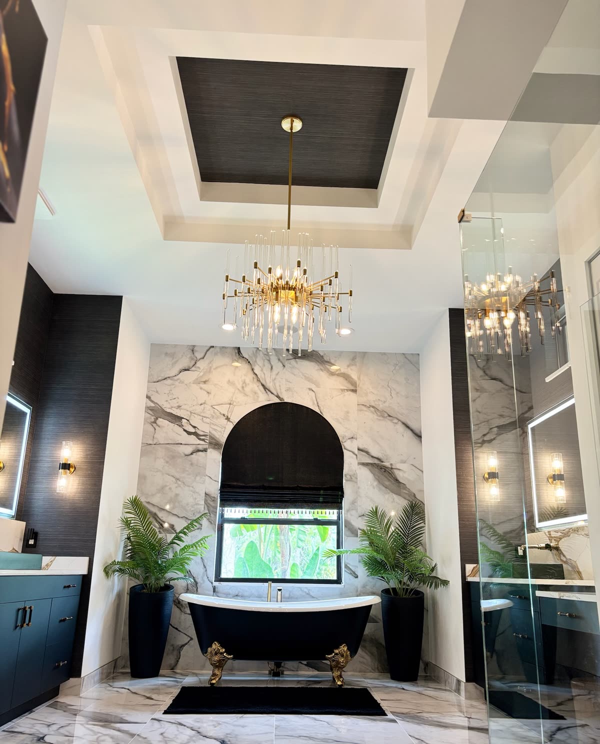 Luxury primary bathroom remodel in Wellington with a black clawfoot tub, a book-matched marble feature wall, navy double vanities, backlit mirrors and a brass chandelier, built by H&H Signature Renovations
